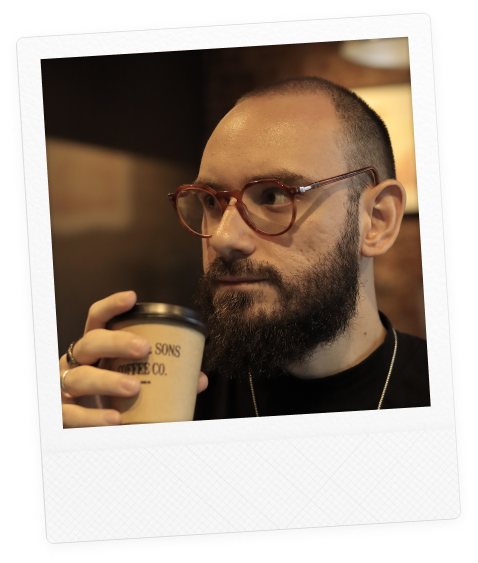 Fotografia de Mateus Villain: Homem de barba cheia e cabeça raspada, usando óculos de armação arredondada, segura um copo de café com tampa enquanto olha para o lado. A foto tem estilo de polaroid, com fundo desfocado em ambiente interno.